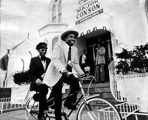 Major Coxson rides a tandem bicycle with his chauffeur, Quinzelle Champagne, in front of Coxson's campaign headquarters in 1972 (his fleet of vehicles had been seized by the IRS).